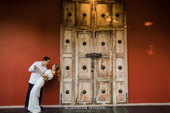 woman in long white dress and man in a guayabera playfully posing next to a rustic wooden door and a red wall